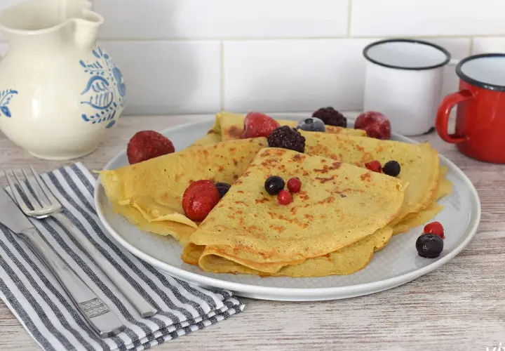 Deliciosos crepes de avena doblados y cubiertos con bayas congeladas y frescas. Servidos en un plato blanco con cubiertos y una jarra de cerámica al fondo.