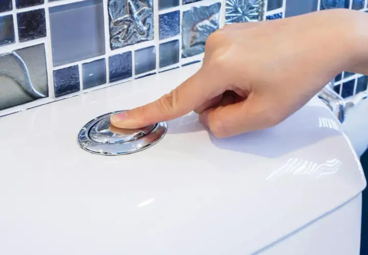 Dłoń naciska chromowany przycisk spłuczki WC na białej ceramice, w tle mozaika.