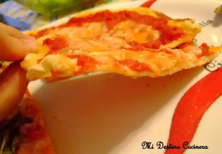 Una mano sostiene una rebanada de pizza con queso derretido y salsa de tomate, sobre un plato decorado con pimientos rojos. La masa, hecha con semola de trigo, luce dorada y crujiente.