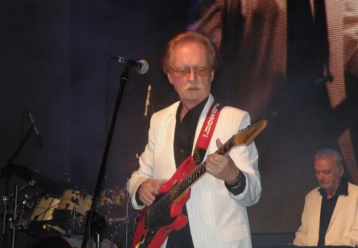 Muzyk w białej marynarce gra na czerwonej gitarze. Na scenie widać też perkusję i klawisze.