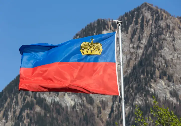 Flaga Liechtensteinu powiewa na tle gór. W jej centrum widnieje złota korona, symbol tego kraju i jego waluty.