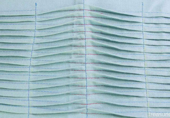 Close-up of light blue fabric with rows of pintucks, creating a textured, pleated effect. Red and blue threads highlight the stitching.
