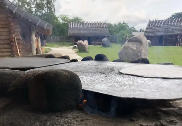 Ognisko pod płaskim kamieniem, na którym piecze się podpłomyk. W tle drewniane chaty i zielony dziedziniec osady średniowiecznej Sławutowo.