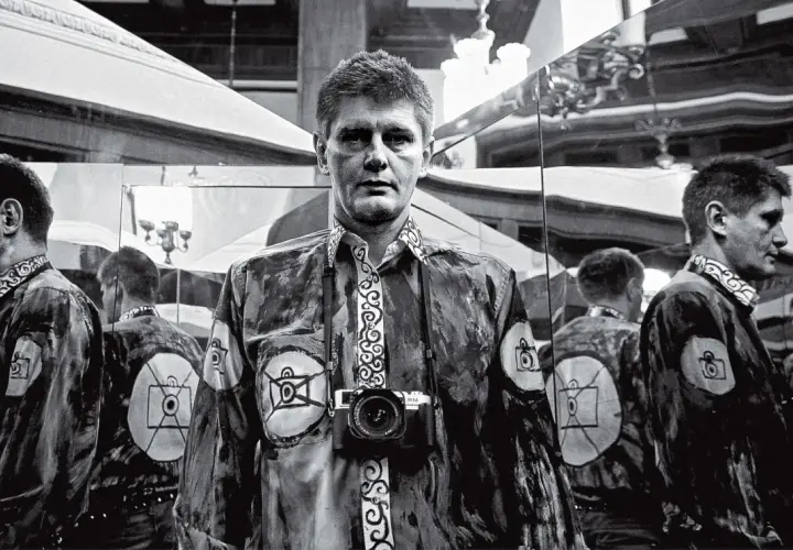 Krzysztof Miller w lustrzanym odbiciu, z aparatem na szyi, w koszuli z motywem oka i aparatu.