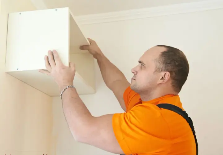 Un homme en orange est en train de fixer un meuble lourd au mur. Il positionne soigneusement l'étagère avant de la visser.