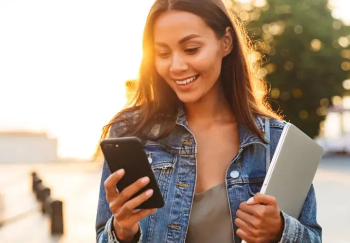 Uśmiechnięta dziewczyna w dżinsowej kurtce czyta na telefonie dwuznaczne teksty do dziewczyny, trzymając laptopa.