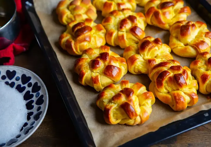Nybakade zeina saffransbullar på en plåt, gyllene och inbjudande. En skål med socker och en röd duk anas i bakgrunden.
