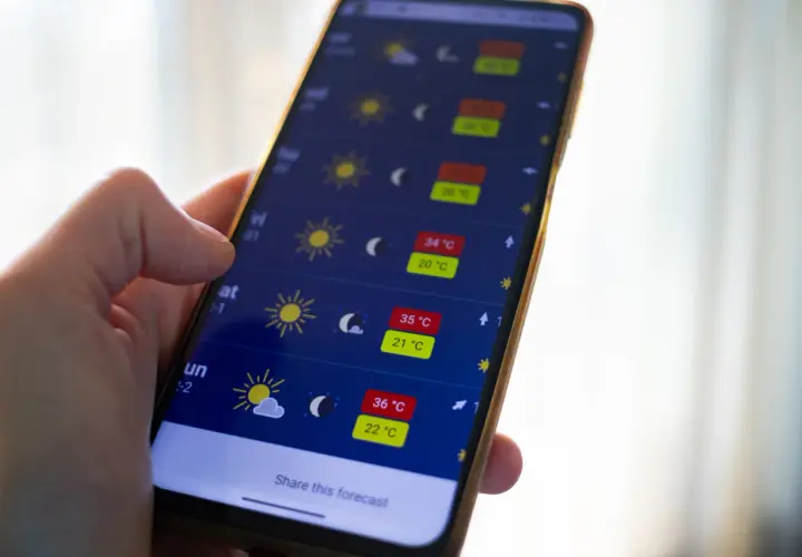 Dłoń trzyma smartfon z aplikacją pogoda, pokazującą wysokie temperatury i słoneczną aurę.