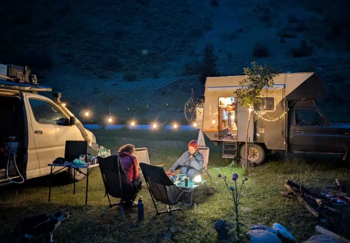 Soirée paisible autour d'un feu de camp, deux personnes assises près d'un **van aménagé électrique** illuminé par des guirlandes lumineuses.