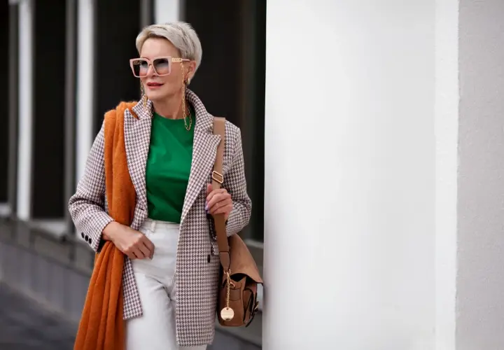 Elegancka kobieta po 60. w stylowej marynarce w kratkę, zielonej bluzce i białych spodniach. Pomarańczowy szal dodaje jej uroku.