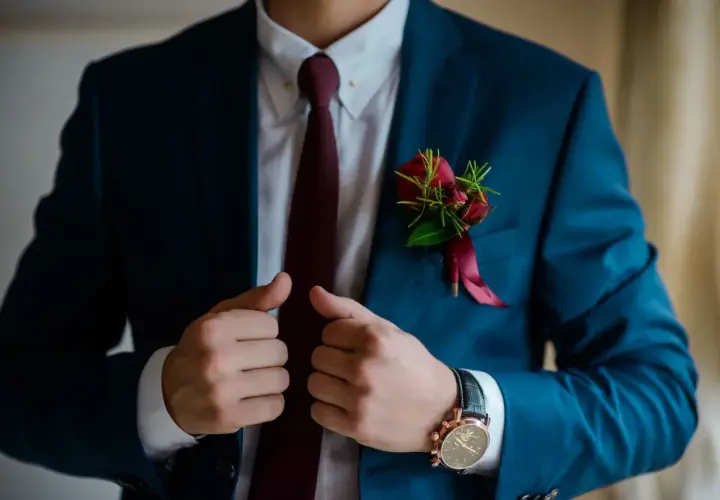 Elegancki mężczyzna w granatowym garniturze, białej koszuli i bordowym krawacie. Na klapie marynarki ozdobna butonierka z czerwonych róż.