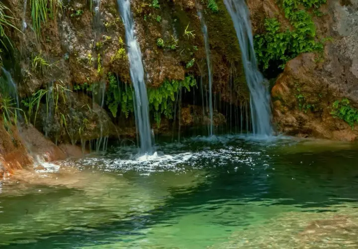 Cascada de la Osera, con agua cayendo sobre rocas cubiertas de musgo y vegetación exuberante, formando un estanque de aguas cristalinas.