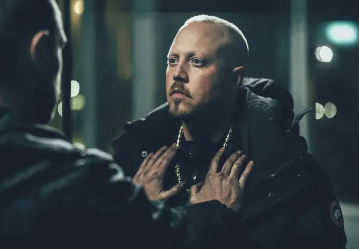 En dramatisk scen från en sebbe stakset film. Två män möts i mörkret, en med guldhalsband, den andra med händerna på hans bröst.