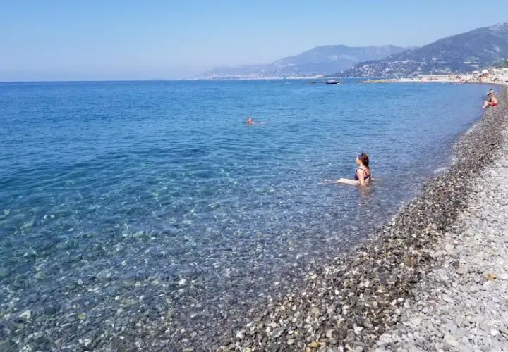 Une femme se baigne dans les eaux claires de la plage de galets de Ventimiglia, avec des montagnes en arrière-plan.