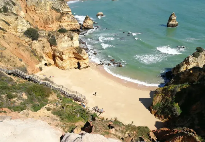 Acantilados dorados y mar turquesa en las **islas de Portugal**. Escaleras de madera descienden a una playa de arena dorada, con formaciones rocosas emergiendo del agua.