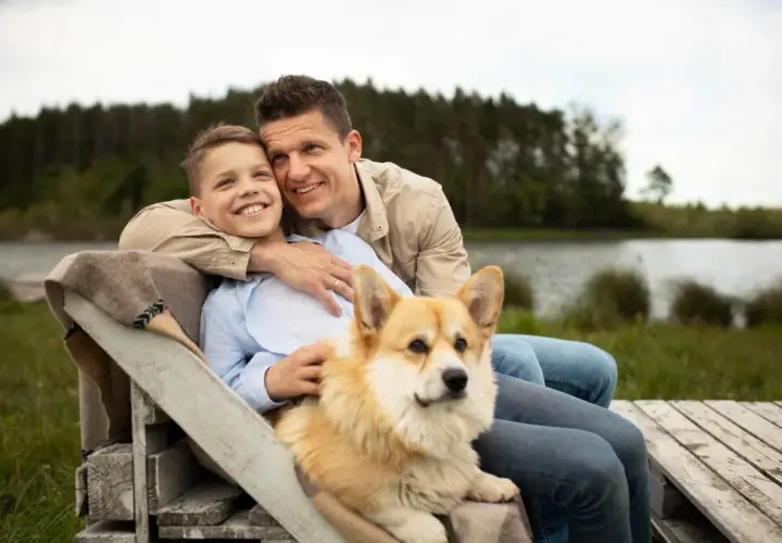 Ojciec i syn siedzą przy jeziorze z psem. Corgi kładzie się na kolanach chłopca, bo czuje się bezpiecznie i kochany.