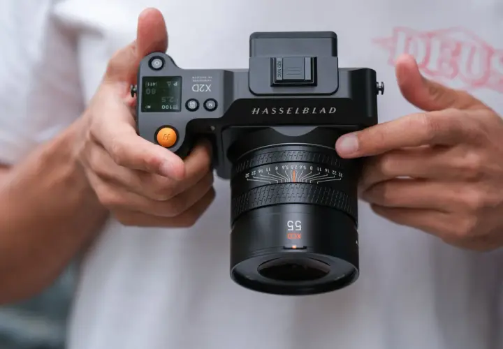 Dłonie trzymają aparat Hasselblad X2D 100C z obiektywem XCD 55mm. Widoczne detale aparatu i obiektywu.