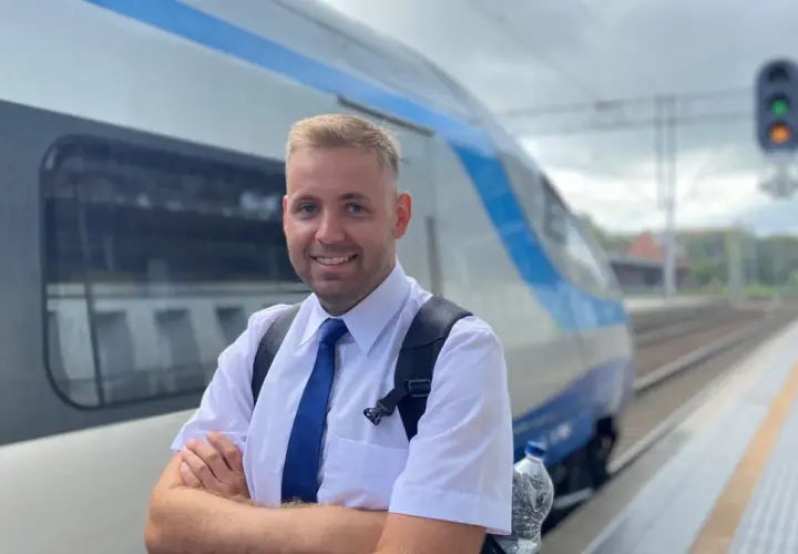 Uśmiechnięty mężczyzna w białej koszuli i niebieskim krawacie, gotowy do pracy. To on prowadzi pociąg, który stoi obok na torach.