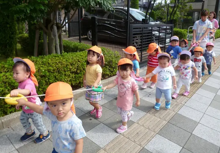 Un gruppo di bambini in Giappone, con cappellini arancioni, cammina in fila tenendo una corda.