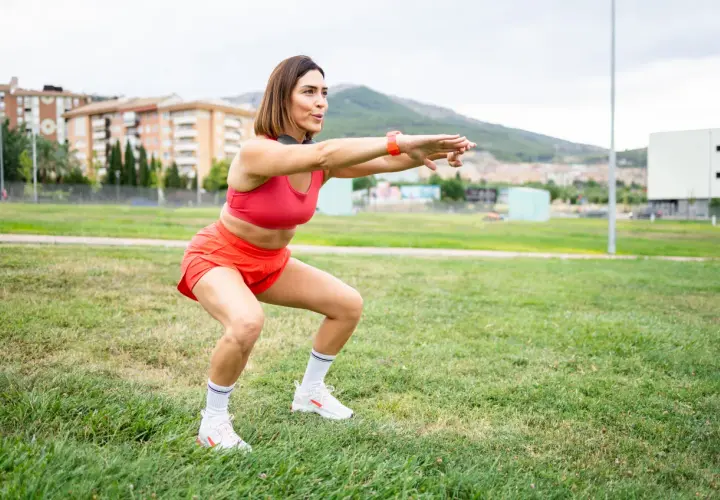 Kobieta w czerwonym stroju sportowym wykonuje przysiady na trawie. To część jej treningu, która pomaga jej odpowiedzieć na pytanie "kim jestem?".