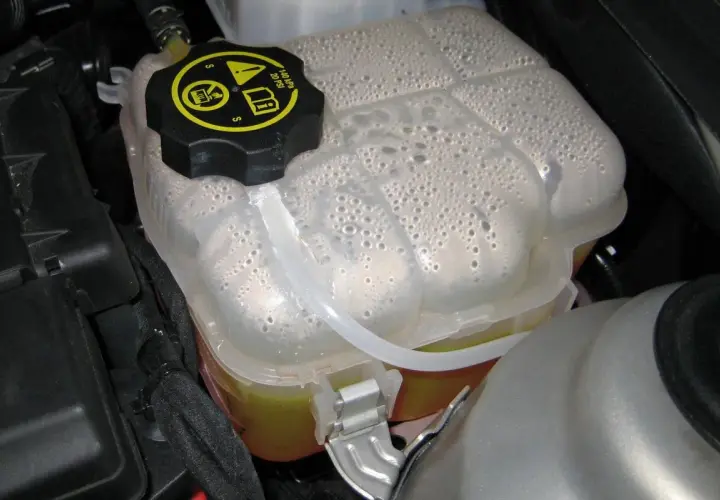 Depósito de expansión del coche con agua y burbujas. El tapón negro tiene símbolos de advertencia.