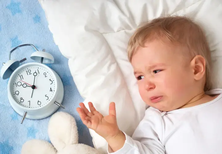 Płacz 1,5 rocznego dziecka, które nie chce spać. Obok budzik i pluszaki.