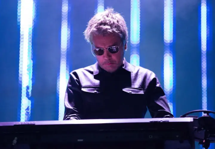 Jean Michel Jarre w Polsce, w okularach przeciwsłonecznych, gra na syntezatorze na tle niebieskich świateł.