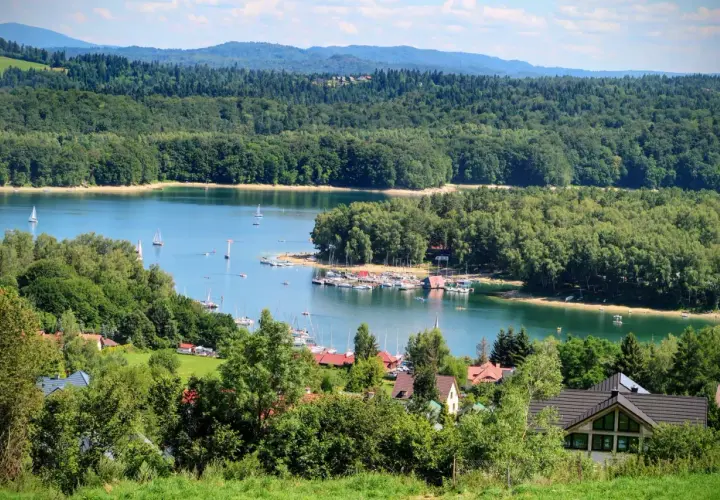 Malowniczy widok na jezioro z żaglówkami i lasem. Widać też domki letniskowe. Czy w Solinie można się kąpać? Tak, to idealne miejsce na wypoczynek!
