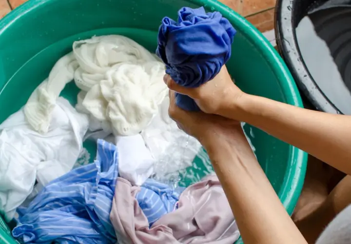 Mains lavant du linge dans une bassine verte. Un vêtement décoloré par le soleil, blanc et bleu, baigne dans l'eau.