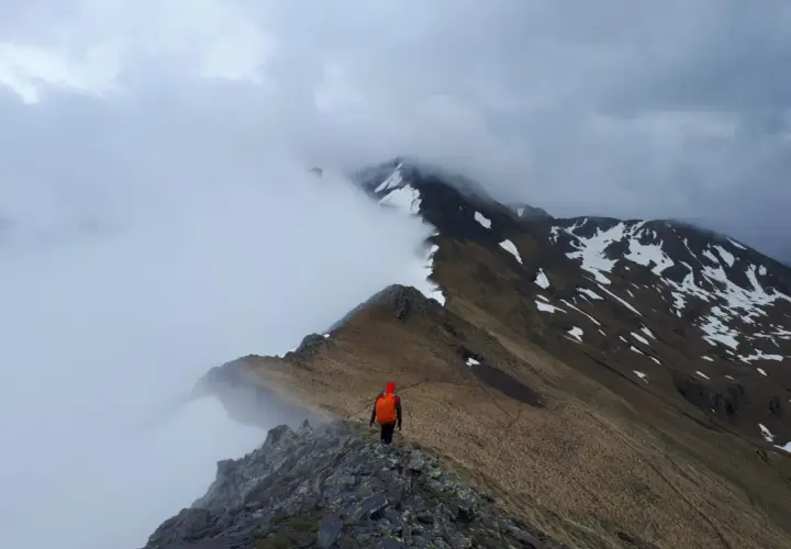 Trekking w Pirenejach. Samotny turysta w pomarańczowym plecaku idzie skalistą granią, otoczony mgłą i ośnieżonymi szczytami.