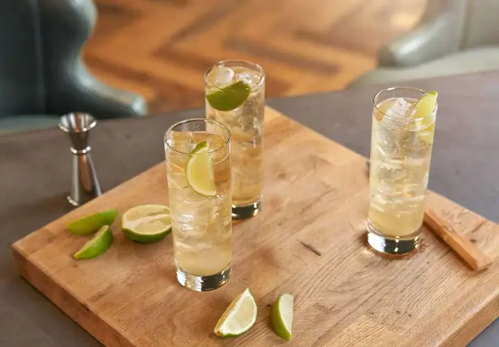 Trzy orzeźwiające whisky ze spritem, udekorowane plasterkami limonki, stoją na drewnianej desce. Obok leżą świeże kawałki limonki.
