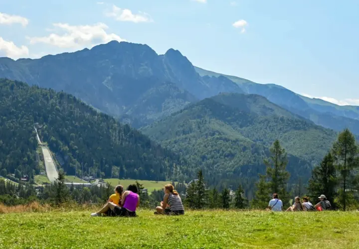 Ludzie odpoczywają na trawie, podziwiając panoramę Tatr z widoczną skocznią narciarską Wielka Krokiew.