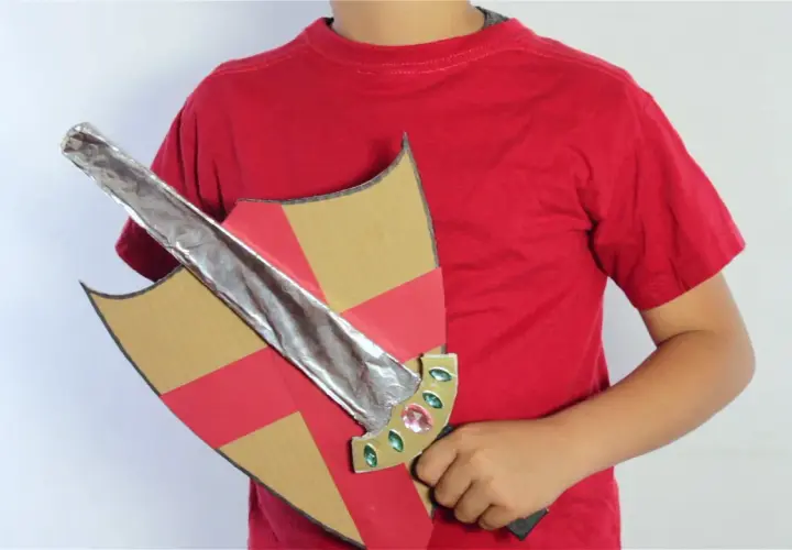 Un niño con camiseta roja sostiene un escudo de cartón con detalles rojos y dorados y una espada de cartón plateada, lista para la aventura.