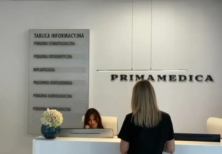Recepcja kliniki PRIMAMEDICA z tablicą informacyjną i pracownicą.