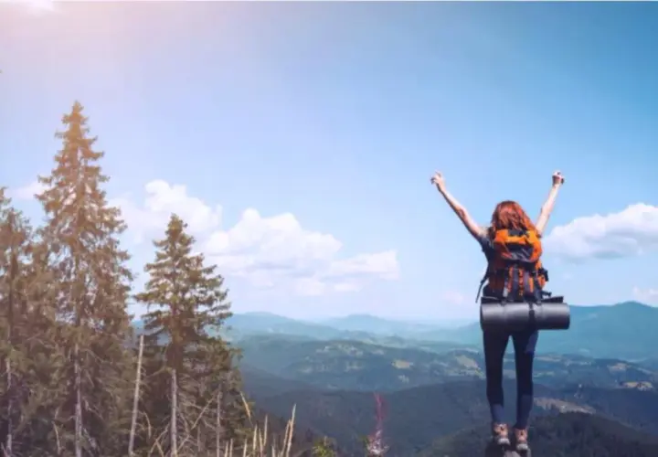 Donna con zaino in cima a una montagna, braccia alzate, esprime il bisogno di viaggiare psicologia.