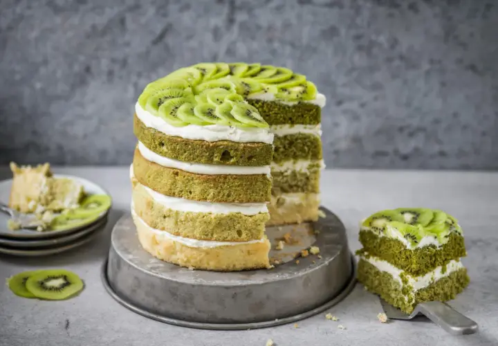 Eine mehrstöckige Matcha Torte mit Kiwi-Scheiben und Sahne. Ein Stück wurde bereits auf einem Teller serviert.