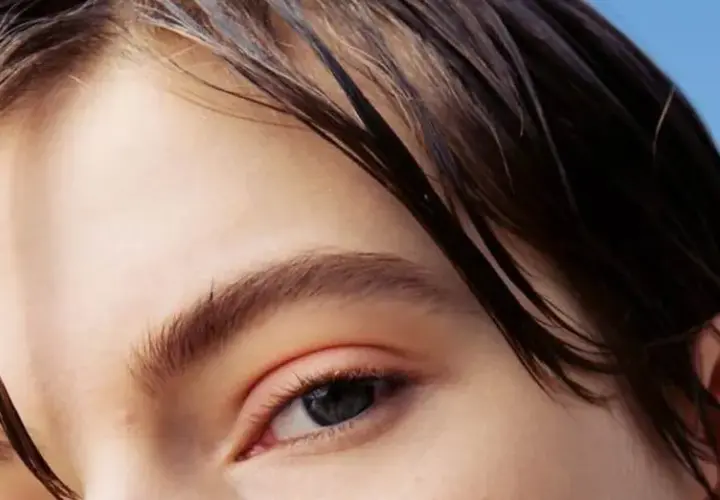 Primer plano de un ojo y cabello oscuro y mojado sobre fondo azul claro.