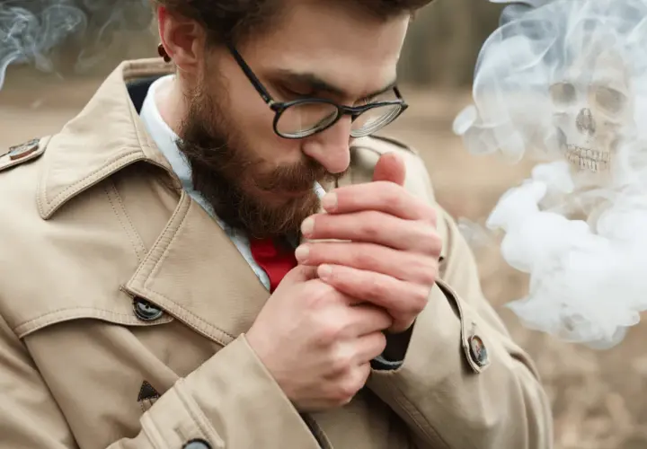 Hombre con barba enciende cigarrillo. El humo forma una calavera, advirtiendo sobre la alergia al humo del tabaco.