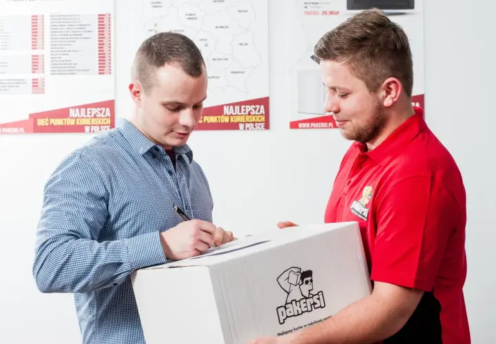 Listonosz w czerwonej koszulce przekazuje paczkę mężczyźnie w kratę, który podpisuje dokument.