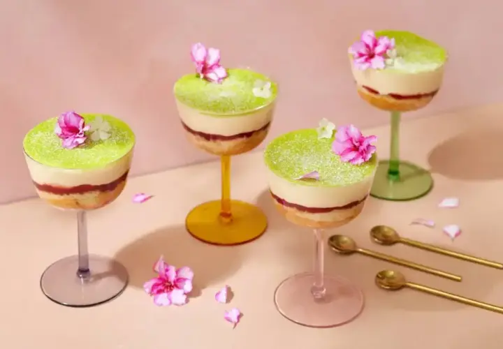 Vackra dessert i glas med lager av kaka, sylt och grön kräm, toppad med blommor. Guldfärgade skedar ligger bredvid.