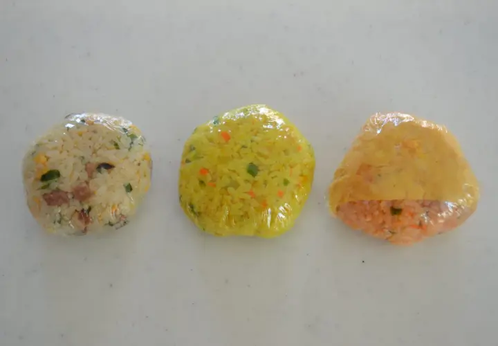 Three plastic-wrapped onigiri sit in a row. The left is white rice with green and brown bits. The middle is yellow rice with orange and green flecks. The right is orange rice with a yellow wrapper. Can you freeze onigiri?