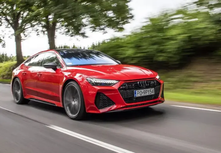 Czerwone Audi RS7 pędzi po drodze, otoczone zielenią drzew.