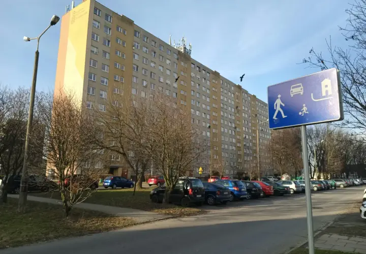 Długi blok mieszkalny, parking z samochodami i znak informujący o możliwości wykupu mieszkania spółdzielczego.