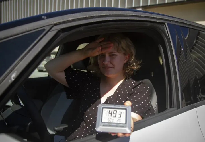 Kvinna i bil svettas och håller upp en termometer som visar 49,3 grader. Det är så varmt att bilen blir inte varm.