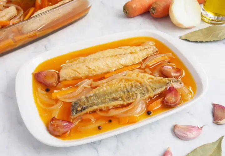 Dos filetes de caballa en escabeche, con zanahoria, cebolla y ajos, en una bandeja blanca.