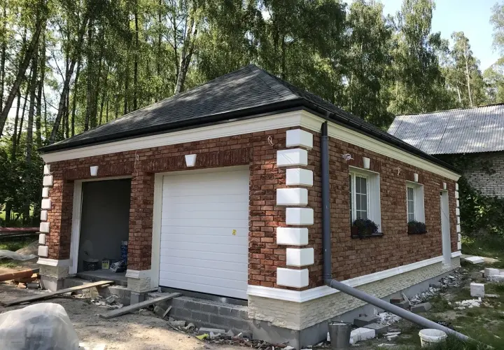 Nowy garaż z cegły, z białym gzymsem i ozdobnymi pilastrami, otoczony zielenią.