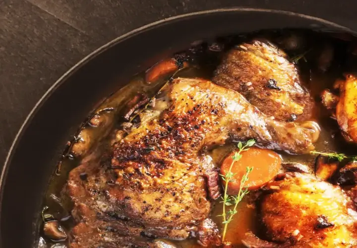 Un plat de coq au vin mijoté, avec des morceaux de poulet dorés, des carottes et des champignons. Le vin rouge utilisé pour la cuisson est essentiel pour ce plat.