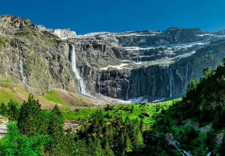 Cascadas impresionantes caen por acantilados rocosos cerca del pueblo de Gavarnie. Un sendero serpentea entre árboles verdes y prados exuberantes.