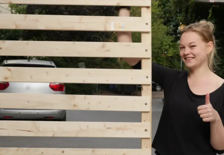 Junge Frau zeigt Daumen hoch neben selbstgebautem Lattenrost. Ein Auto ist im Hintergrund zu sehen.