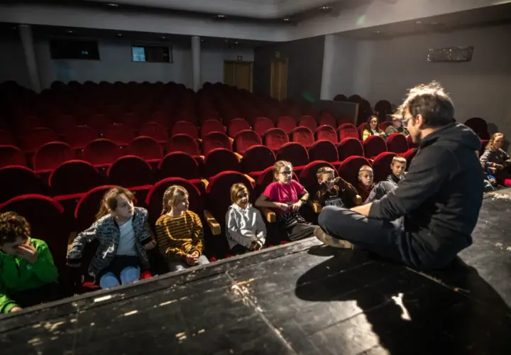 Dzieci słuchają opowieści na scenie, siedząc przed rzędem czerwonych foteli. To część repertuaru teatru Zagłębia.